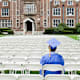 College graduate student wearing a blue cap and gown sits alone in an empty outdoor auditorium before his graduation ceremony.