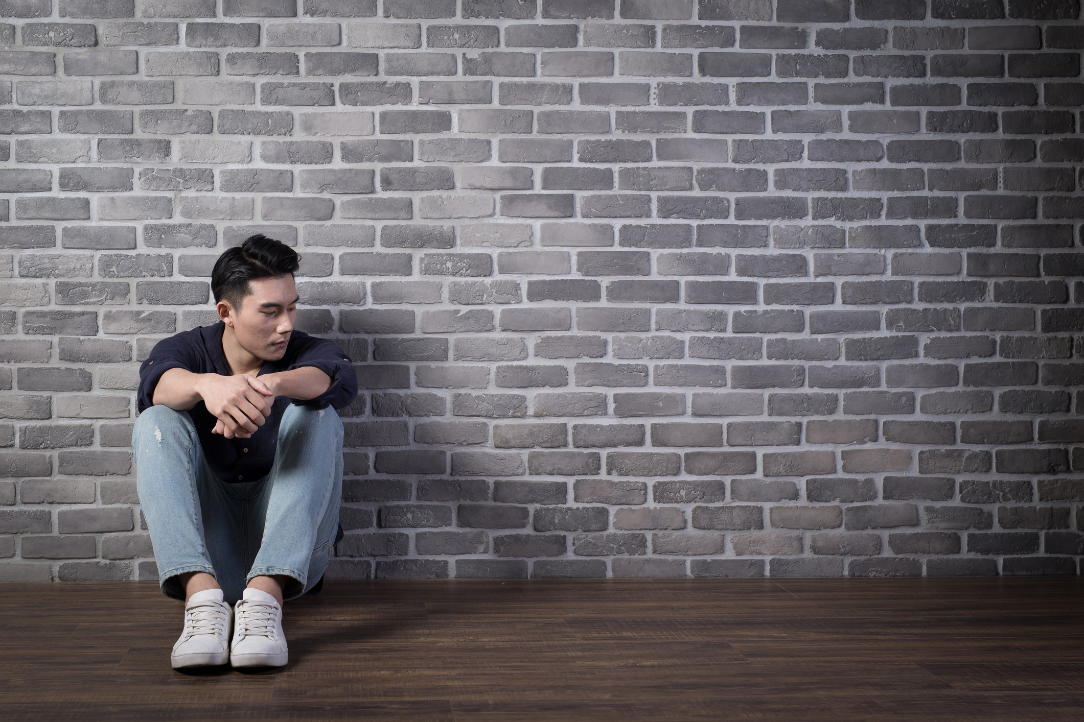 A young man sits with his back against a brick wall, looking down in a worried state.