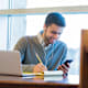 Student checking college application deadlines on his mobile phone while working on college application forms for his laptop.