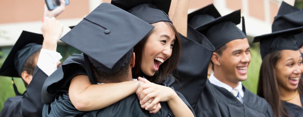 A group of college students dressed in caps and gowns congratulate and hug each other on graduation day.