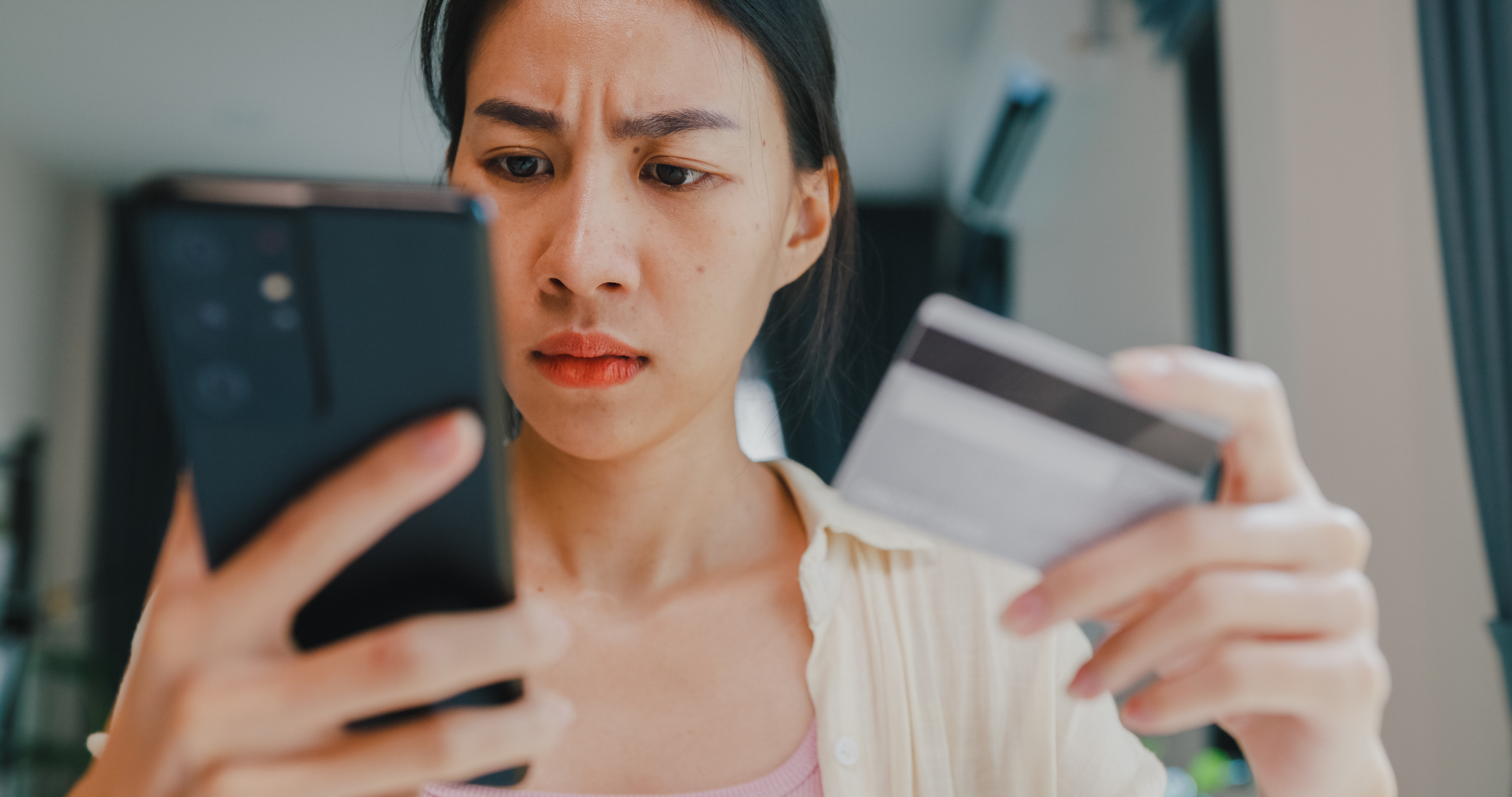 Asian American female college student using her credit card to pay her student loan bill. She is looking at her mobile phone, looking shocked at the amount she has to pay.