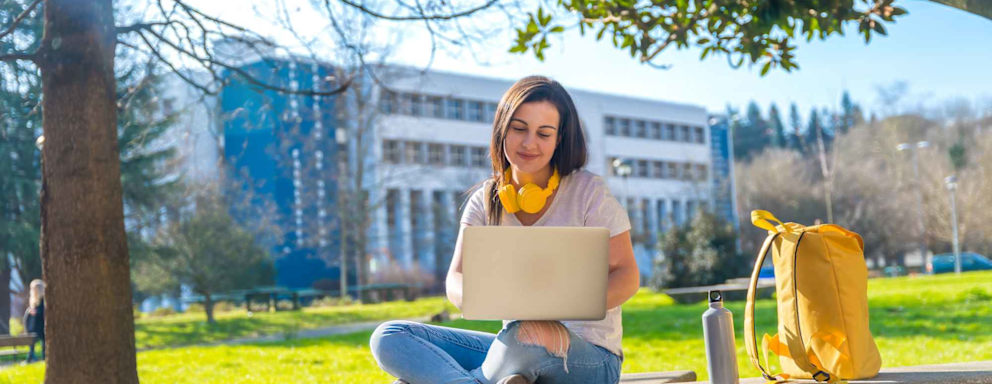 Caucasian female college student sitting on a bench on a college campus quad outside. She is doing homework on her laptop.