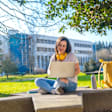 Caucasian female college student sitting on a bench on a college campus quad outside. She is doing homework on her laptop.