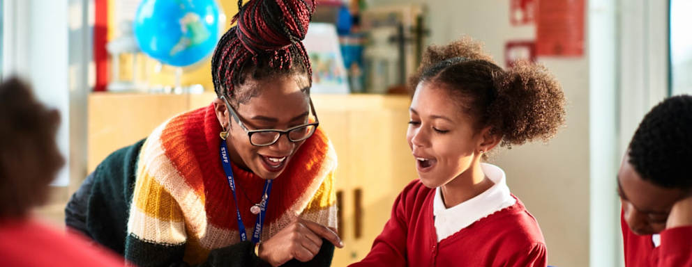 A Black female elementary school teacher helps a student in her classroom with homework.