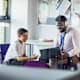A male secondary school student sitting in an office in the school, having a meeting with his teacher. The teacher is showing him something on his laptop while they both smile.