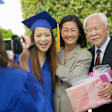 A first-generation Asian American female student poses with her mother and grandfather at her graduation ceremony. She is wearing a blue cap and gown. A friend is videotaping her and her relatives in celebration of her achievements.