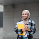 A first-generation Black female student wearing a hijab scarf and casual Western coat and jeans walks to class on campus. She is holding a pair of notebooks in her right arm and a cup of coffee in her left. She is smiling and looking happy.