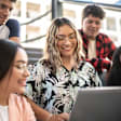 A Latina college student sits with her friends in a university classroom. She is smiling while showing them something on her laptop.