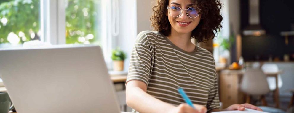 A Caucasian female college student sitting at her dining room table at home. She has her laptop open and is taking notes for class.