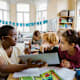 Black female teacher holding a tablet device to help explain a concept to an elementary-aged schoolboy and schoolgirl at their classroom desks.