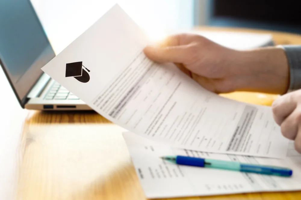 A close-up of a hand holding a financial aid award letter on a desk with a laptop in the background.